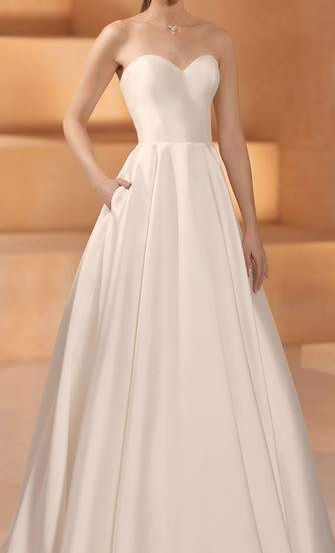 Eine Frau in einem trägerlosen weißen Hochzeitskleid steht drinnen und posiert mit einer Hand auf der Hüfte. Der Hintergrund zeigt eine beige Treppe.