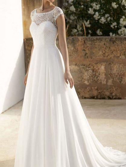 Eine Frau in einem weißen Hochzeitskleid mit Spitzenbesatz steht auf einem Betonboden, mit einer Steinmauer und weißen Blumen im Hintergrund.