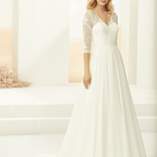 Eine Frau mit blonden Haaren steht drinnen und trägt ein weißes V-Neck-Hochzeitskleid mit Spitzenärmeln. Das Kleid hat einen fließenden Rock und ist vor einem neutralen Hintergrund platziert.