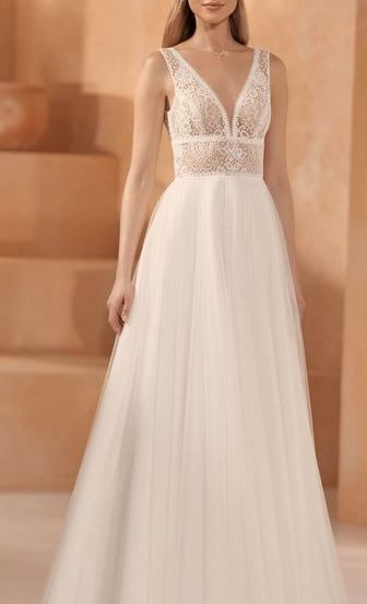 Eine Frau in einem weißen Hochzeitskleid mit Spitzendetails steht in einem Raum mit beigem Hintergrund und Treppen.