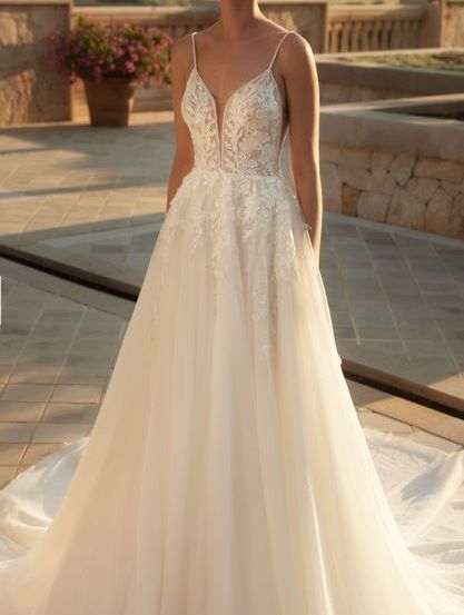 Eine Frau in einem weißen Hochzeitskleid mit aufwendiger Spitze steht auf einer Terrasse mit einer Steinmauer und Blick auf Berge und Blumen.