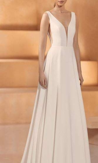 Eine Frau in einem weißen, V-Ausschnitt-Hochzeitskleid steht auf einer Bühne, trägt eine Halskette und hält eine Clutch.