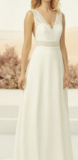 Eine Frau mit blondem Haar steht in einem weißen Hochzeitskleid mit Spitzendetails auf einer weißen Plattform. Der Hintergrund ist verschwommen mit rosa Pflanzen.