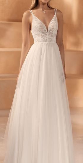 Eine Frau in einem weißen Hochzeitskleid mit aufwendigen Spitzendetails steht in einem warmen, neutral getönten Raum.