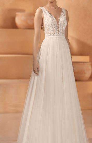 Eine Frau in einem weißen, mit Spitze verzierten Hochzeitskleid steht auf einer Bühne mit beigefarbenen Stufen und einem beigefarbenen Hintergrund.