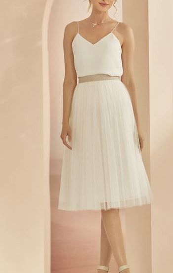 Eine Frau steht in einem Raum und trägt ein weißes ärmelloses Kleid mit einem plissierten Rock und einer Halskette. Sie posiert mit gekreuzten Beinen.