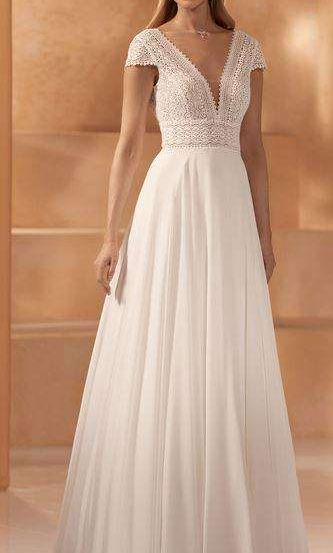 Eine Frau in einem weißen, mit Spitze verzierten Hochzeitskleid mit V-Ausschnitt und Falten steht vor einer pfirsichfarbenen Wand.