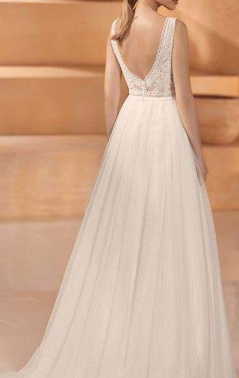 Eine Frau mit einem Pferdeschwanz steht vor einem beigen Hintergrund. Sie trägt ein weißes, elegantes Hochzeitskleid mit V-Ausschnitt und Spitzendetails.