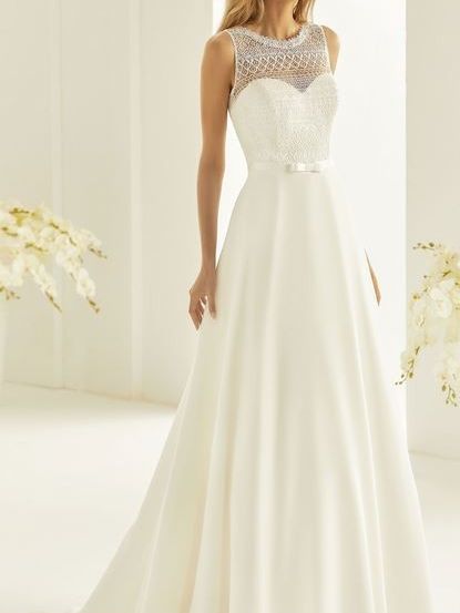 Eine Frau in einem weißen Hochzeitskleid steht vor einer weißen Wand, mit einem Strauß weißer Blumen im Hintergrund.