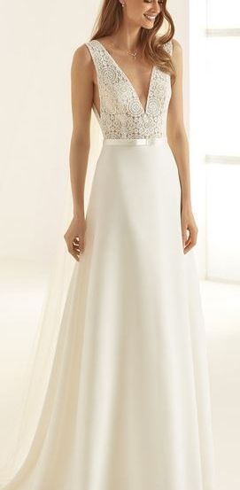 Eine Frau in einem weißen Hochzeitskleid mit Spitze und einem silbernen Gürtel, das gegen einen weißen Hintergrund steht und lächelnd in die Kamera schaut.