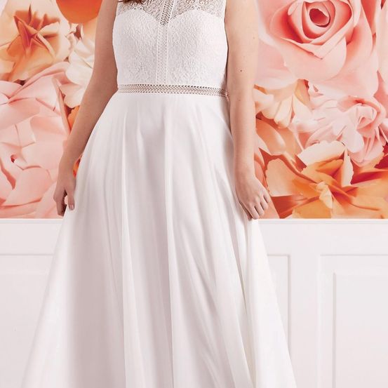 Eine Frau steht vor einem Hintergrund großer rosa Blumen und trägt ein weißes Hochzeitskleid mit Spitzendetails am Oberteil und einem silbernen Gürtel.