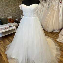 Ein weißes Hochzeitskleid auf einem Mannequin in einem Raum mit Holzboden. Hinter ihm befinden sich weitere Hochzeitskleider an Kleiderständern.