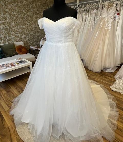Ein weißes Hochzeitskleid auf einem Mannequin in einem Raum mit Holzboden. Hinter ihm befinden sich weitere Hochzeitskleider an Kleiderständern.