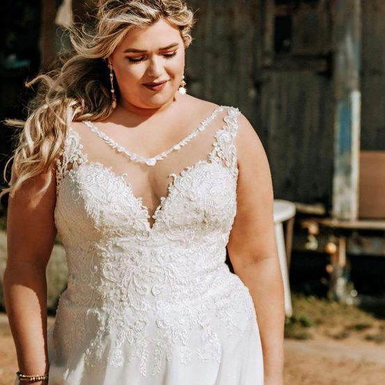 Eine Frau mit blondem Haar, die ein weißes Hochzeitskleid trägt, steht im Freien. Sie lächelt, ihr Kleid hat Spitzendetails und einen V-Ausschnitt. Sie steht vor einem rustikalen Gebäude.