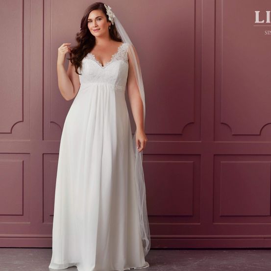 Eine Frau in einem weißen Hochzeitskleid mit langem Schleier steht vor einer rosa Wand. Sie lächelt und scheint für ein Foto zu posieren.