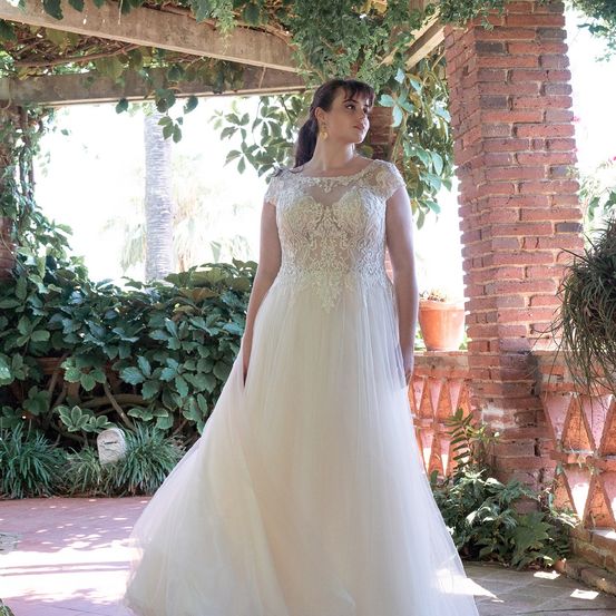 Eine Frau in einem Hochzeitskleid steht auf einer Veranda, umgeben von Grün und einer Ziegelmauer.