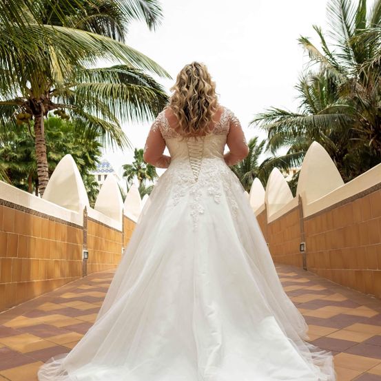 Eine Frau mit blonden Haaren geht einen Gehweg entlang und trägt ein weißes Hochzeitskleid mit aufwendigen Spitzendetails und einem korsettartigen Oberteil. Im Hintergrund befinden sich Palmen und ein Gebäude.