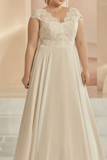 Eine Frau in einem cremefarbenen, langen Hochzeitskleid mit Spitze am Oberteil, steht auf einem weißen Boden.