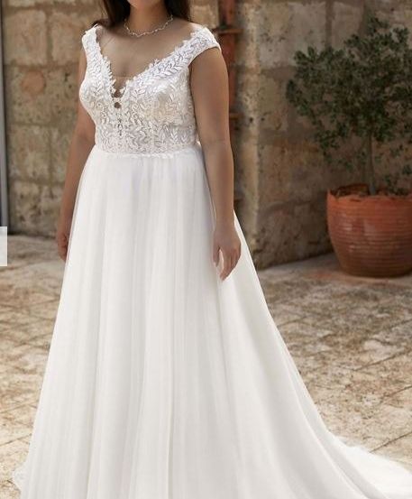 Eine lächelnde Frau in einem weißen Hochzeitskleid steht vor einer Steinmauer und einem Topf mit einer Pflanze.