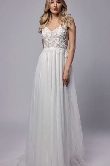 Eine Frau mit blonden Haaren trägt ein weißes Hochzeitskleid mit aufwendigen Spitzendetails am Oberteil. Das Kleid fällt elegant bis zum Boden.