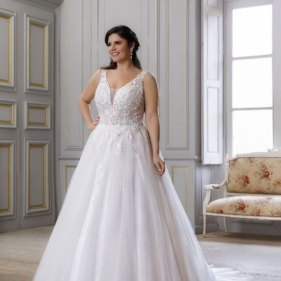 Eine Frau in einem weißen Hochzeitskleid steht in einem Raum mit einer weißen Wand und einem weißen Stuhl.