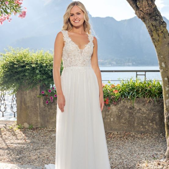 Eine lächelnde blonde Frau steht auf einem Kiesweg und trägt ein weißes Hochzeitskleid. Hinter ihr befindet sich eine Steinmauer mit Pflanzen und Blumen. Im Hintergrund sind Berge und ein See zu sehen.