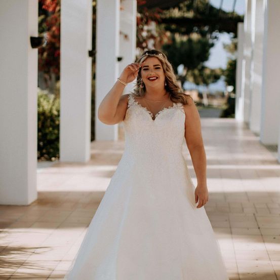 Eine Frau in einem weißen Brautkleid steht auf einem Gehweg, lächelt und posiert für ein Foto. Sie trägt eine Krone und Schmuck, und der Hintergrund zeigt einen Garten mit Pflanzen und Bäumen.