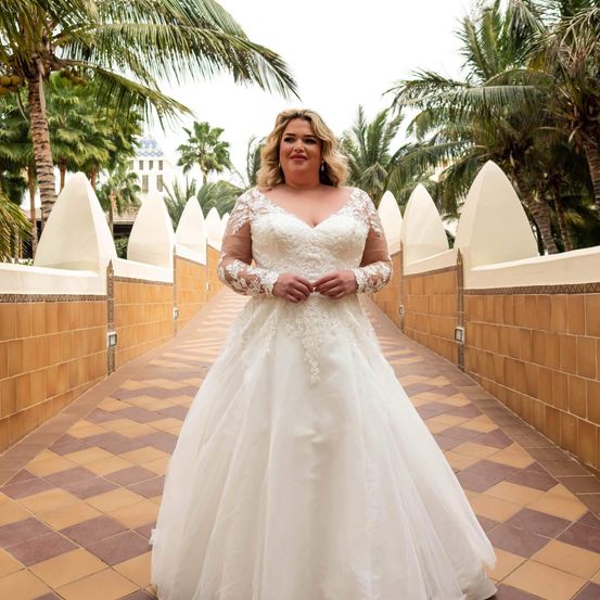 Eine lächelnde Frau in einem Hochzeitskleid steht auf einem gefliesten Weg mit Palmen und Bogen im Hintergrund.