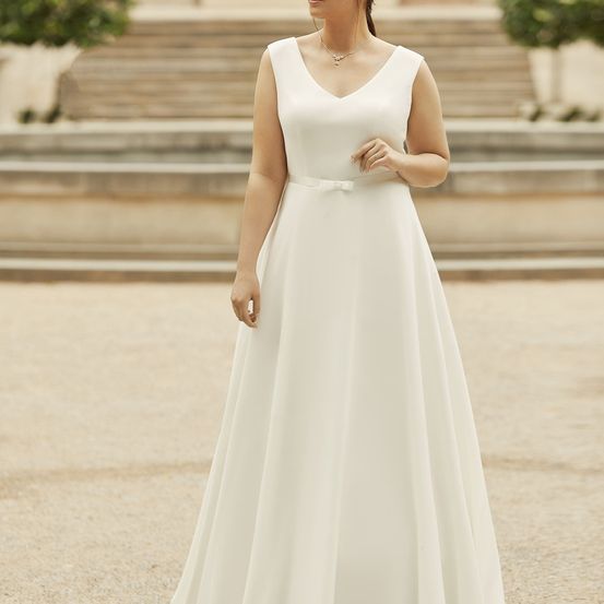 Eine Frau in einem weißen, ärmellosen Kleid steht auf einem Betonboden mit Stufen hinter ihr. Sie trägt eine Halskette und einen Gürtel. Die Szene ist im Freien.