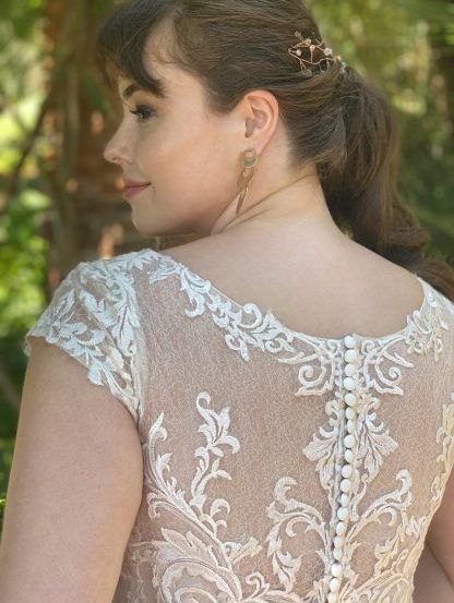 Eine Frau mit braunen Haaren in einem Pferdeschwanz steht nach links gewandt und trägt ein Brautkleid aus Spitze mit komplizierten Mustern.