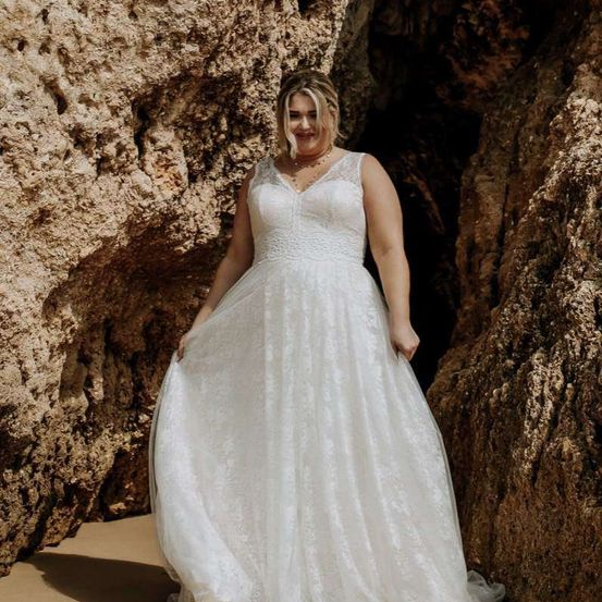 Eine Frau steht in einem weißen Hochzeitskleid am Strand zwischen großen Felsen. Sie lächelt in die Kamera.