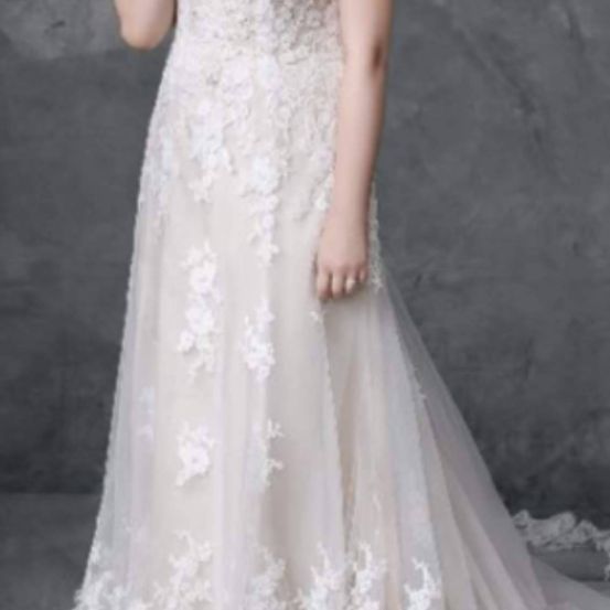 Eine Frau in einem weißen Hochzeitskleid mit komplizierten Spitzenmustern steht vor einem grauen Hintergrund. Das Kleid hat eine taillierte Corsage und einen fließenden Rock.