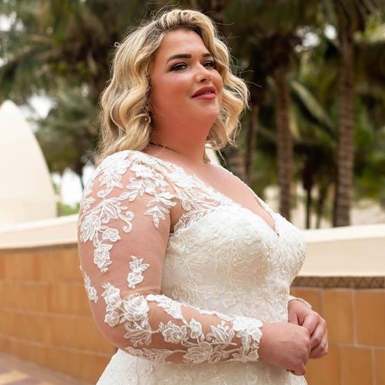 Eine Frau mit blonden Haaren in einem weißen Spitzenhochzeitskleid posiert für ein Foto in einer Außenumgebung mit einer Steinmauer und Palmen im Hintergrund.