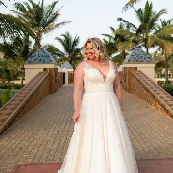 Eine Frau in einem Hochzeitskleid steht auf einer Brücke, lächelt und schaut nach vorne, mit dekorativen Gebäuden und Palmen im Hintergrund.