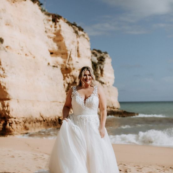 Eine Frau in einem Hochzeitskleid steht auf einem sandigen Strand mit einer Klippe im Hintergrund.