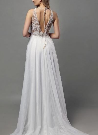 Eine Frau mit blonden Haaren trägt ein weißes Hochzeitskleid mit Rückenausschnitt und langem Schleier in einem grauen Raum.