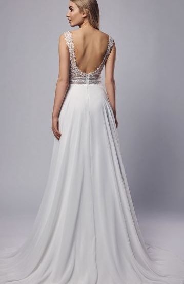 Eine Frau mit blonden Haaren ist von hinten zu sehen, sie trägt ein weißes Hochzeitskleid mit einem aufwendigen Rückendesign. Das Kleid fällt bis zum Boden.