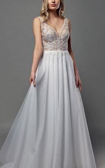 Eine Frau in einem weißen Hochzeitskleid mit Spitzendetails steht vor einem grauen Hintergrund. Das Kleid hat einen V-Ausschnitt und einen fließenden Rock.