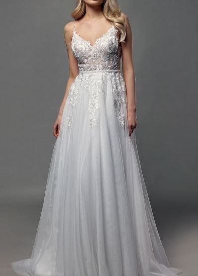 Eine Frau mit langen blonden Haaren posiert in einem weißen Hochzeitskleid mit komplizierten Spitzendetails vor grauem Hintergrund.