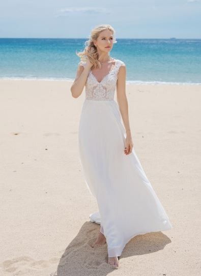 Eine Frau in einem weißen Hochzeitskleid steht am Strand, mit Blick auf das Meer, und hat die Hände auf die Hüften gestemmt.