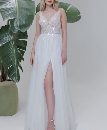 Eine Frau in einem weißen Kleid mit Schlitz posiert neben einer großen grünen Pflanze vor weißem Hintergrund.