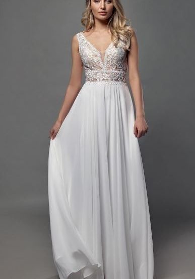 Eine Frau mit blondem Haar steht in einem Studio und trägt ein weißes, fließendes Hochzeitskleid mit Spitzendetails. Sie blickt mit neutralem Ausdruck nach vorne.