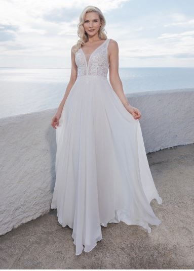 Eine Frau mit blondem Haar steht auf einem Betonbalkon und trägt ein weißes Hochzeitskleid mit V-Ausschnitt und Spitzendetails. Das Kleid hat einen fließenden Rock und sie schaut auf das Meer hinaus.