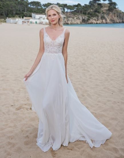Eine Frau steht am Strand in einem weißen Hochzeitskleid und posiert für ein Foto mit einem Felsen und dem Meer im Hintergrund.