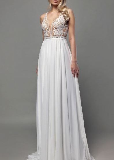 Eine Frau posiert in einem weißen Kleid mit einem mit Spitze verzierten Oberteil und einem bodenlangen Rock vor einem grauen Hintergrund.