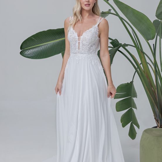 Eine Frau mit blondem Haar posiert in einem weißen Spitzenkleid neben einer großen grünen Pflanze mit breiten Blättern.