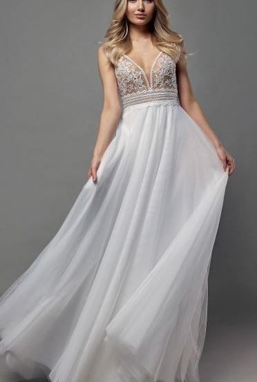 Eine Frau mit blonden Haaren posiert in einem weißen Kleid mit Spitzendetails vor grauem Hintergrund.