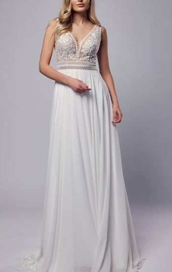 Eine Frau in einem weißen Hochzeitskleid mit aufwendiger Spitzenverzierung am Oberteil steht vor grauem Hintergrund. Das Kleid hat einen V-Ausschnitt und einen fließenden Rock.