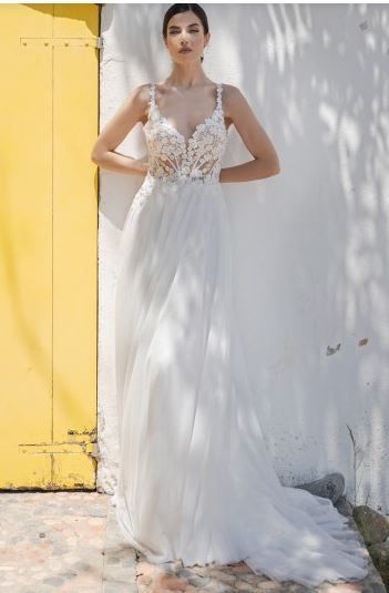 Eine Frau in einem weißen Hochzeitskleid mit Spitzendetails steht vor einer weißen Wand mit einer gelben Tür.