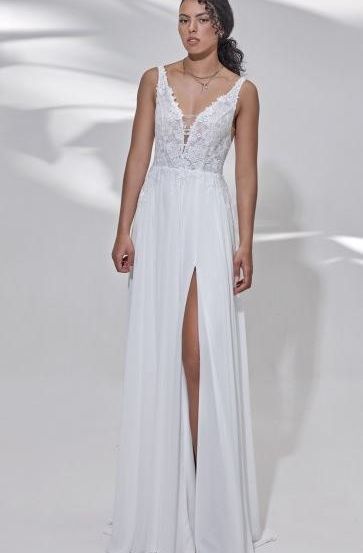 Eine Frau steht in einem weißen Kleid mit tiefem V-Ausschnitt und Spitzendetails, das eine Halskette trägt. Das Kleid hat einen hohen Schlitz auf einer Seite, und sie posiert vor einem weißen Hintergrund.